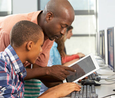 Teacher Helping Boy To Use Digital Tablet In Computer Class Having A Discussion